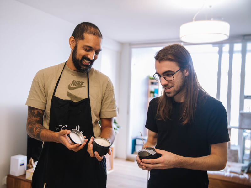 Two men wearing aprons and smiling whilst holding handmade candles.