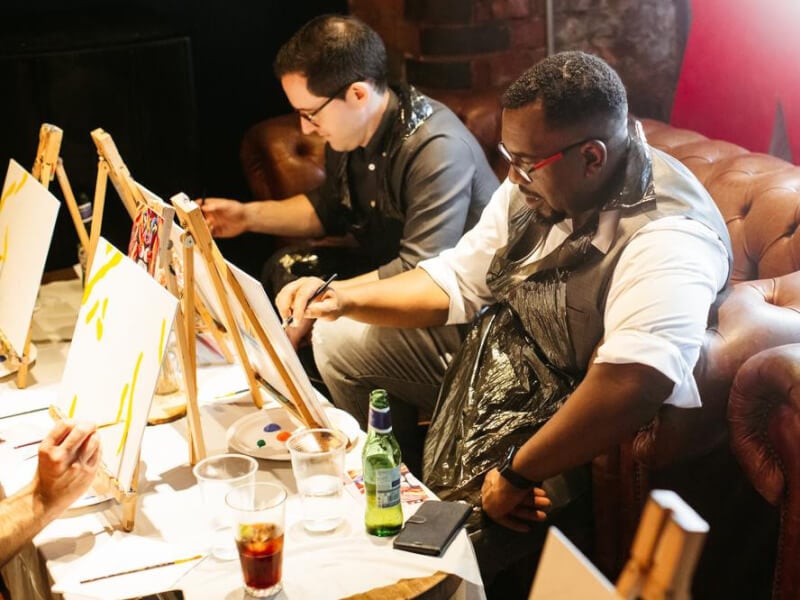 Two men sitting on a leather chesterfield sofa painting on small canvases