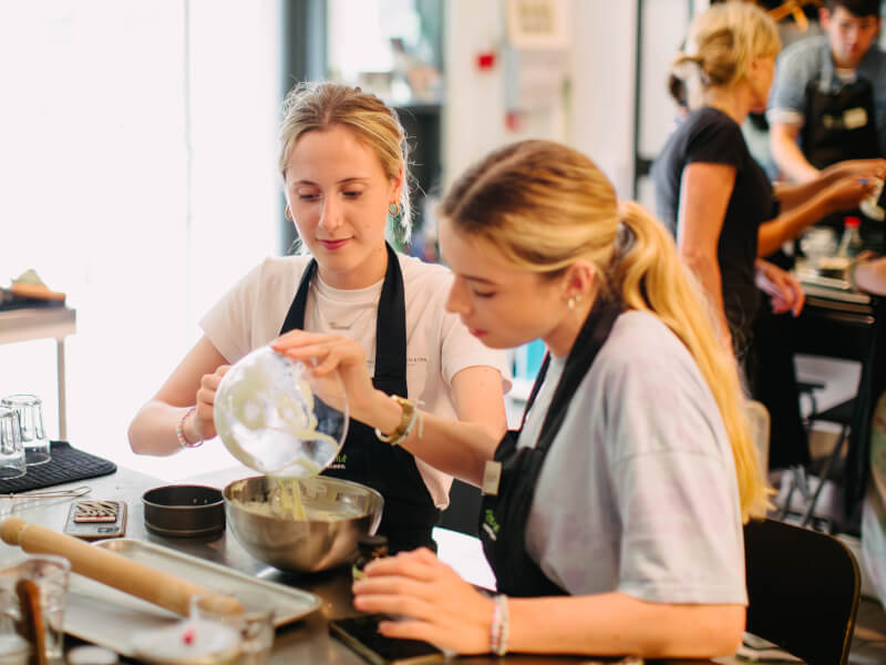 Two women at a cooking class are pouring ingredients from a glass bowl to a metal mixing bowl