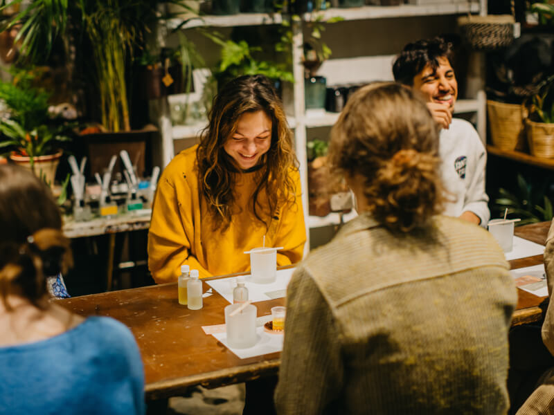 Woman in a yellow knit jumper smiling down at a frosted glass candle with a wick holder and fragrance oils by her side
