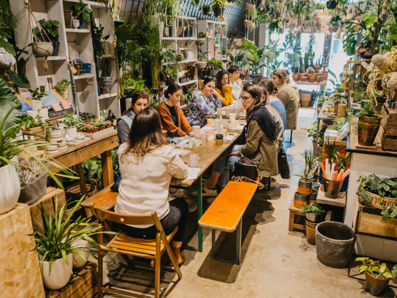 A group of people sit at a table at a candle making class, surrounded by plants