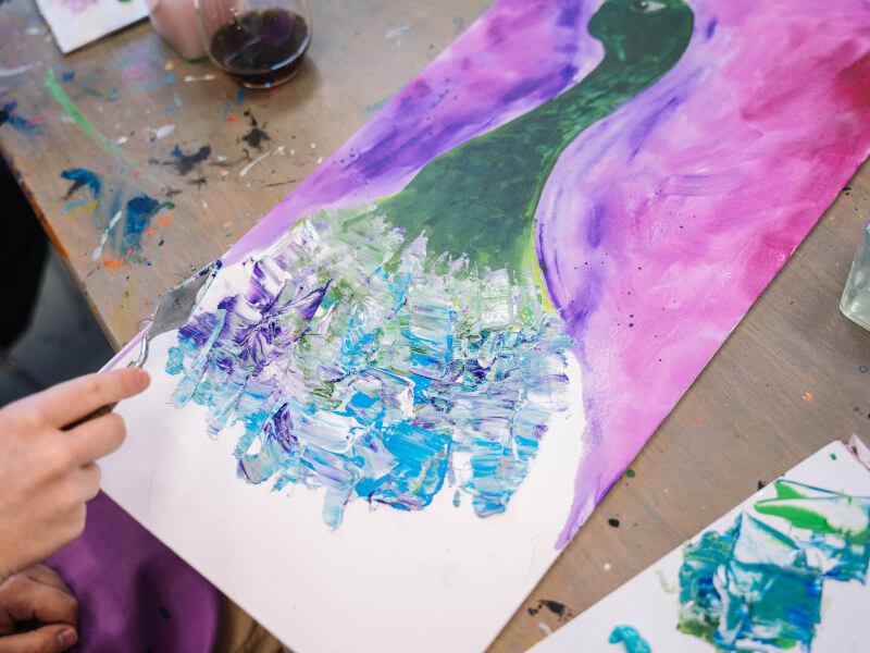 A close up of a person applying blue and purple paint to a canvas with a palette knife