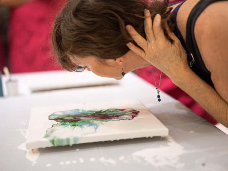 A person is leaning over a canvas and blowing on paint after it's been poured on at a fluid art class