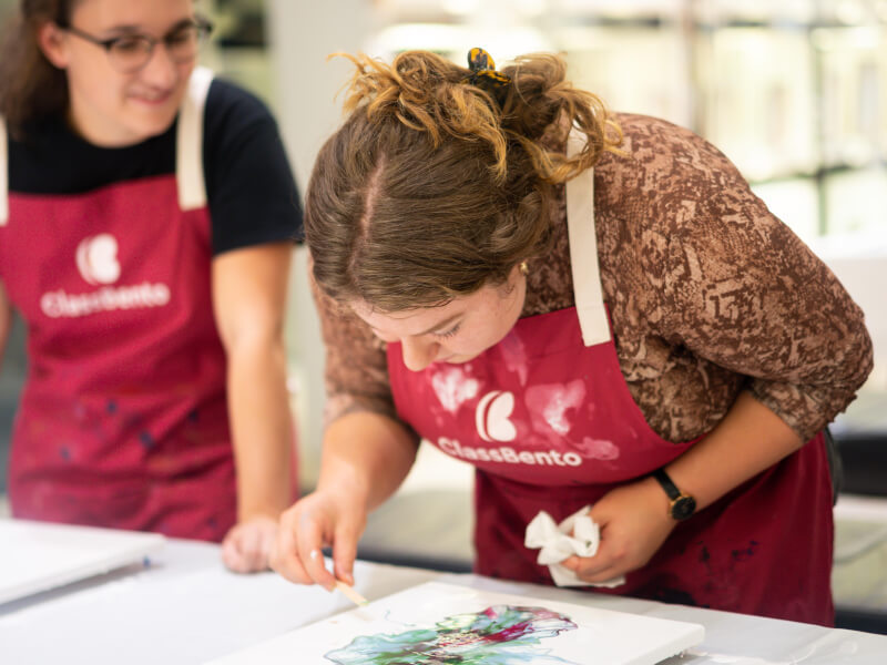 Two women are standing at a table, one is looking closely at a work of art she's making with paint