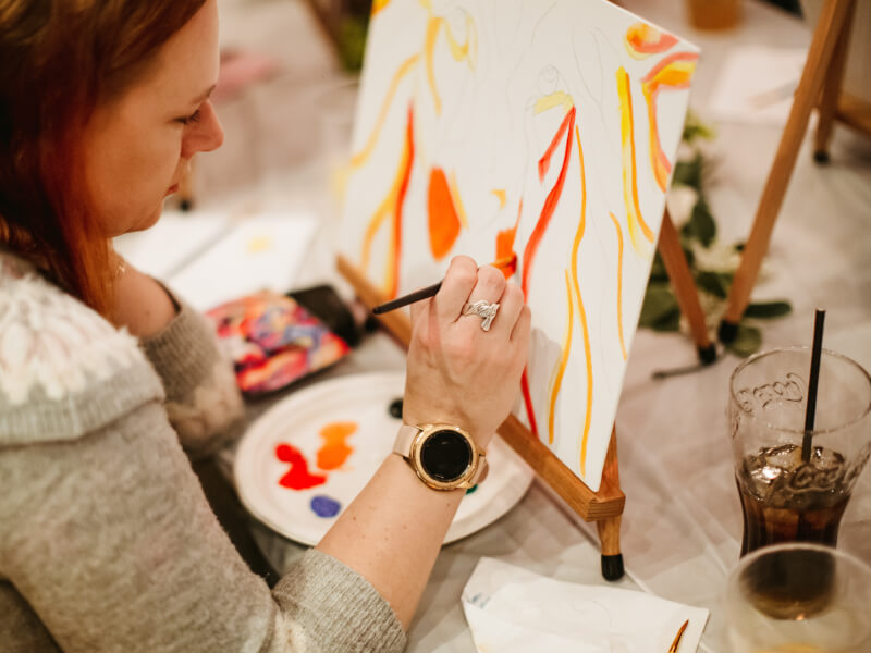 A woman is sitting at an easel on a table, painting a multicoloured lion head on a canvas.