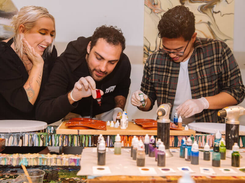 Two men and a woman are standing at a table adding coloured ink to a mixing pallette