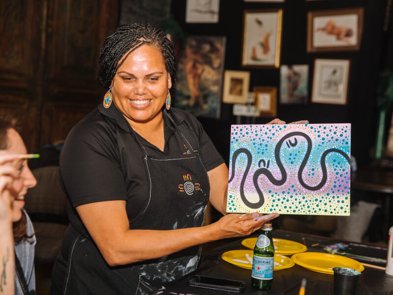 A woman is standing up holding a canvas with a colourful work of art in purple, blue and black.