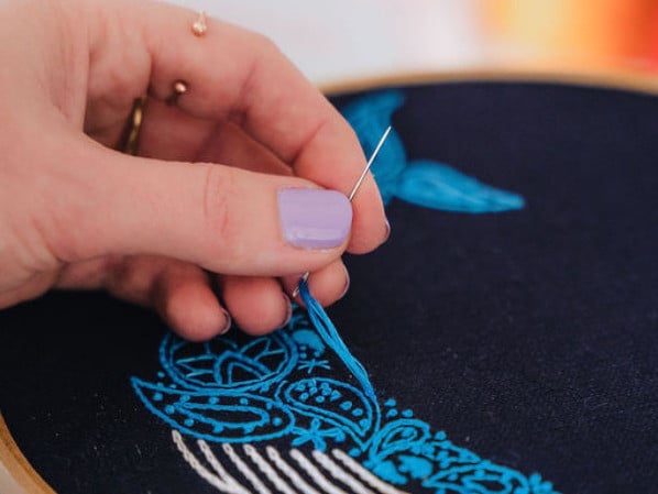 A close up of a person embroidering a blue whale design on black fabric in a hoop