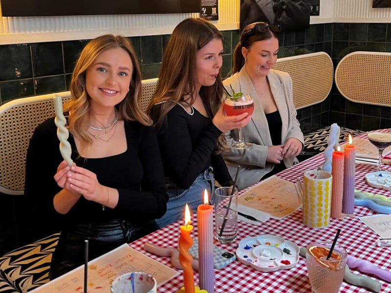 Three women at a Bendy Boy candle making workshop are surrounded by handmade candles and smiling 