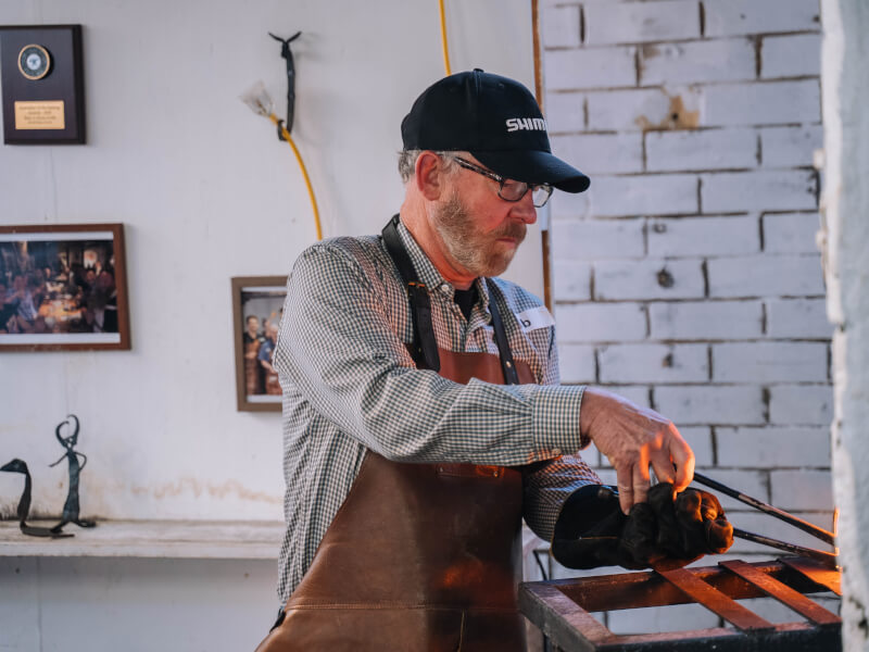 A man is wearing a leather apron and using tools at the fire in a forge