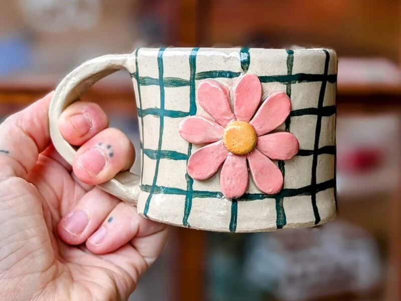 A hand holding a handmade clay mug, decorated with a green grid and pink flower