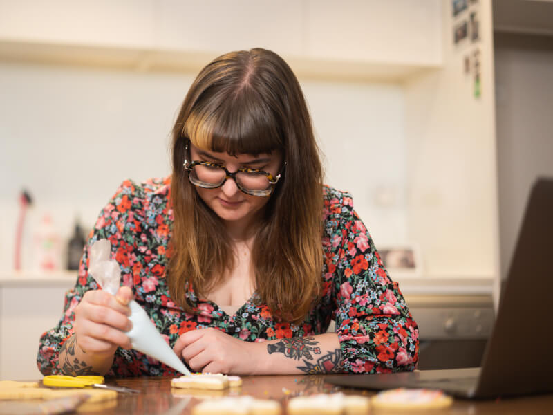 A woman is sitting at a table, using an icing bag and nozzle to ice a biscuit