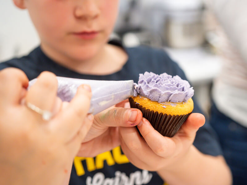 A child is decorating a cupcake in a brown wrapper with purple frosting using a piping bag