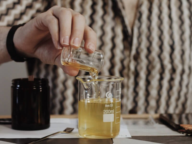 A close up of a person pouring melted wax into a jar from a small glass beaker