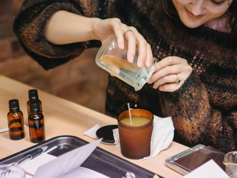 A close up of a woman pouring melted wax into a container with a wick