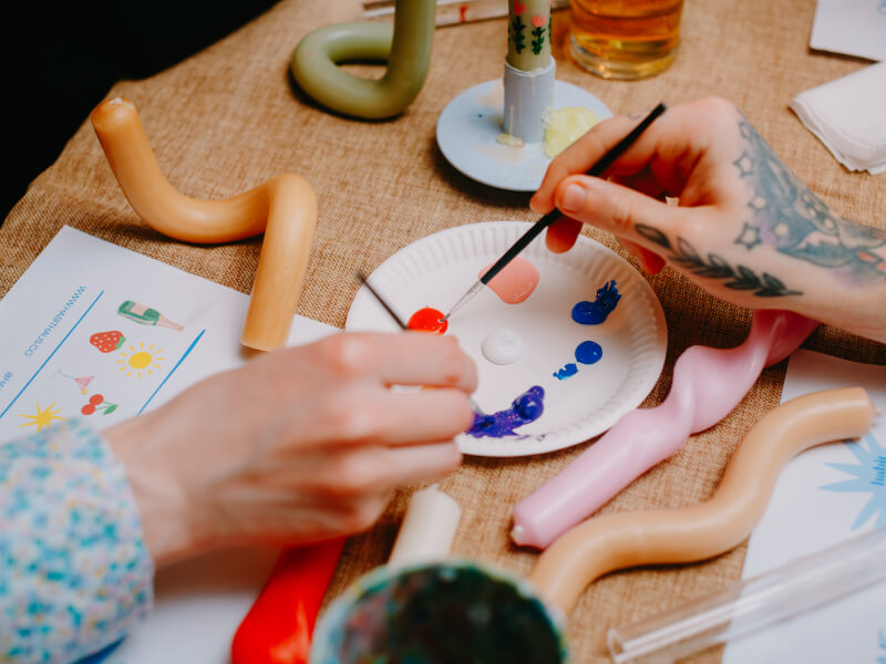 A close up of two peoples hands dipping brushes into coloured paint to paint candles