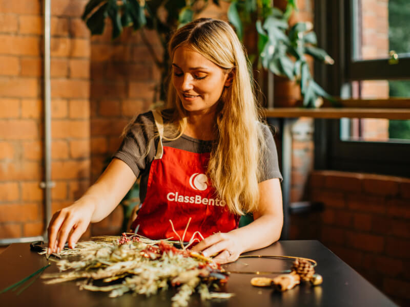 A woman is sitting at a wooden table making a flower wreath from dried flowers
