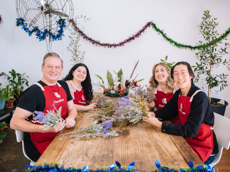 A group of people are sitting at a wooden table with dried purple flowers, holding them in bunches