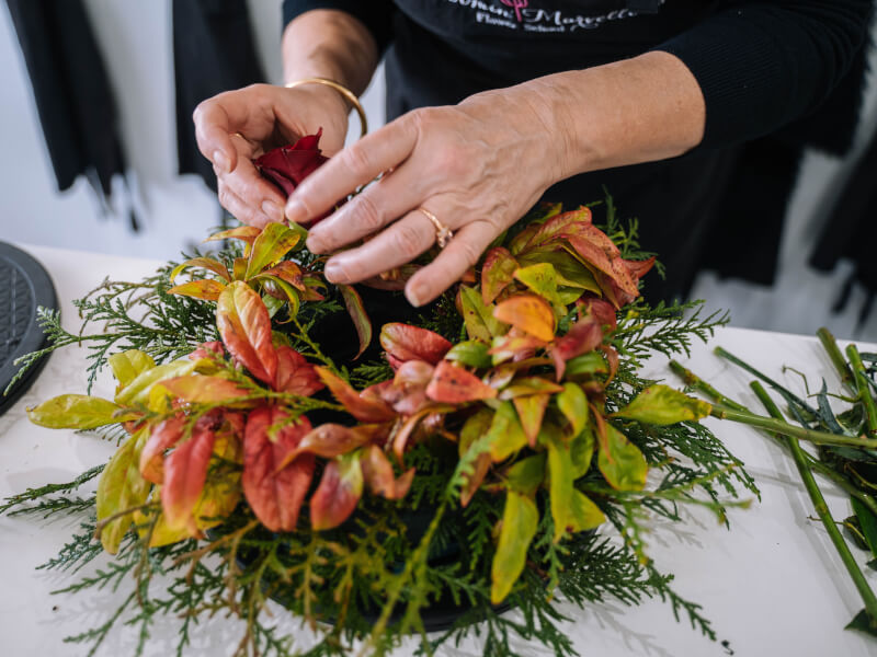 A close up of a person making a large flower wreath from red leaves and green foliage