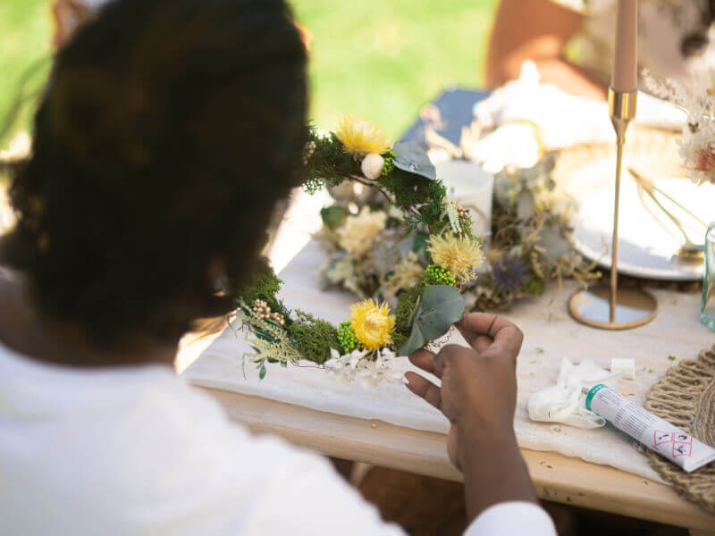 A person at an outdoor flower wreath class is attaching yellow flowers and green foliage to a wreath base