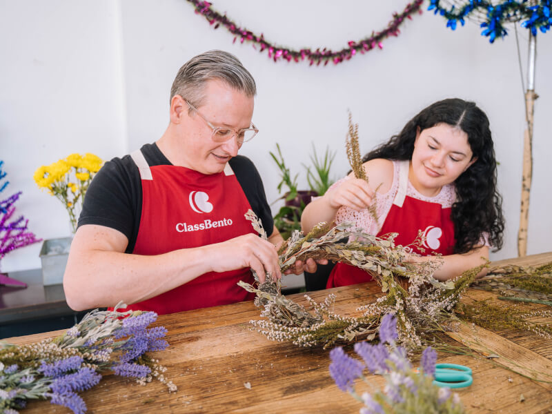 A man and woman are attaching dried flowers to a wreath base and smiling
