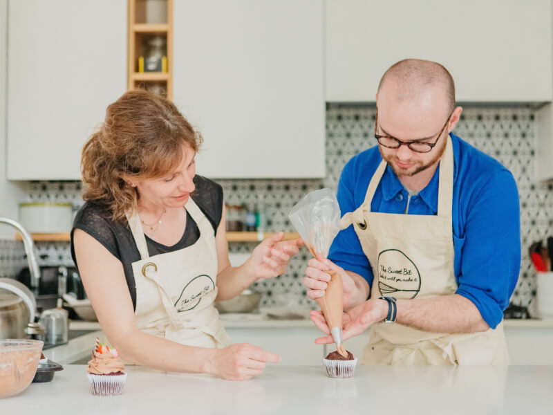 A man and woman are adding chocolate buttercream to the top of a cupcake using a piping bag and nozzle