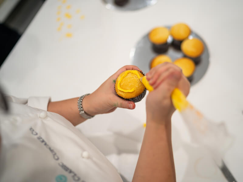 A woman is icing a cupcake with yellow buttercream frosting at a cake decorating class