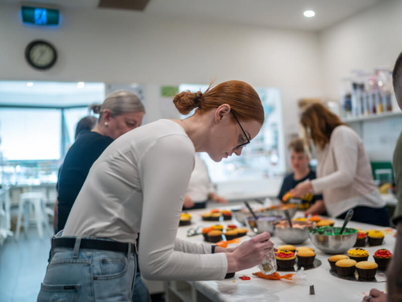 Several people are standing around a table, decorating cupcakes with brightly coloured buttercream frosting