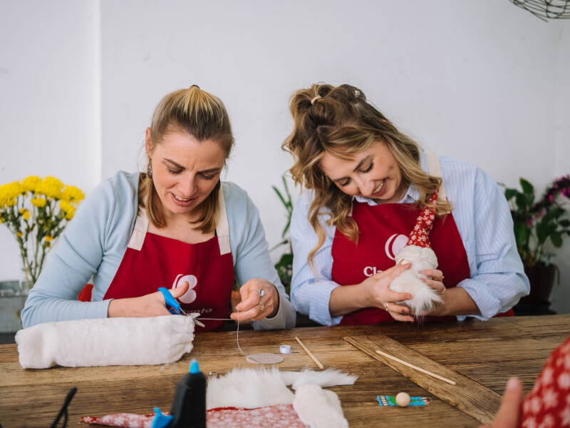 Two women are sitting at a desk making a Santa Claus decoration