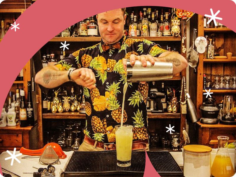 A man stands behind a bar pouring a yellow cocktail from a shaker into a glass filled with ice