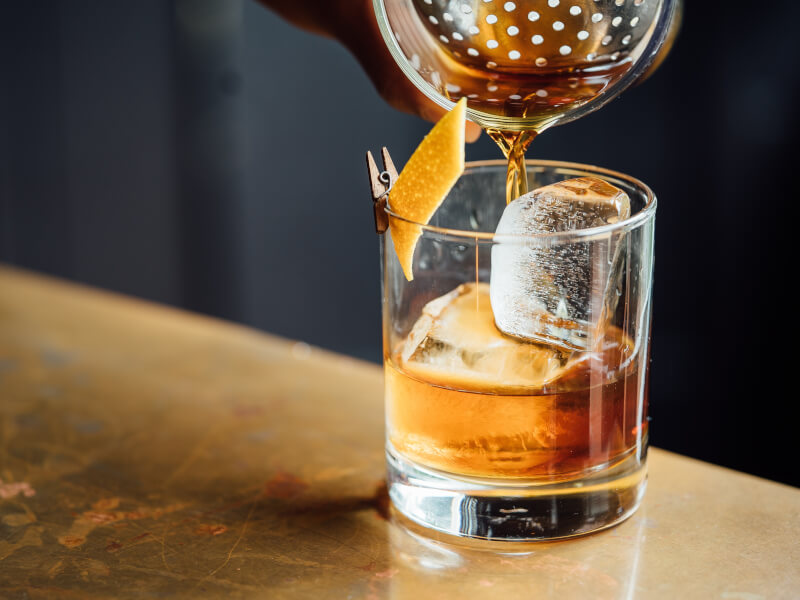 A person pouring whiskey into a glass filled with ice and decorated with lemon