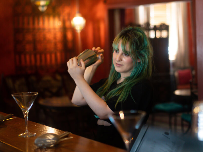 A woman with green hair is shaking a cocktail shaker in both hands
