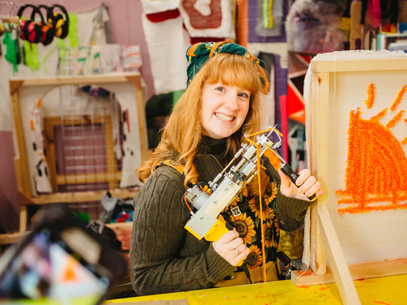 A woman is holding a tufting gun loaded with yellow yarn and standing behind a canvas with a sun design