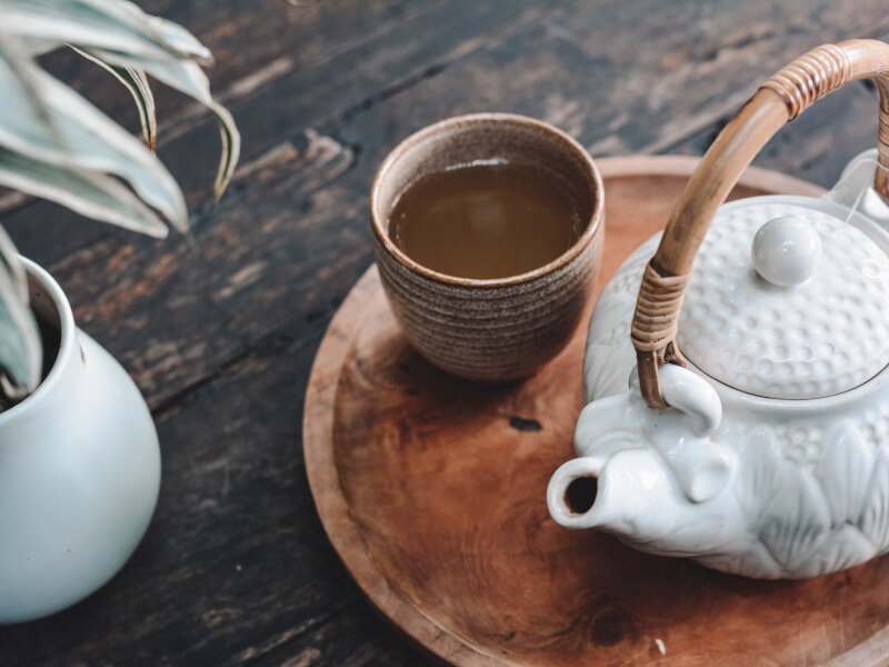 A white teapot and brown cup have been placed on a wooden tray, on a table. There is a plant to the left.