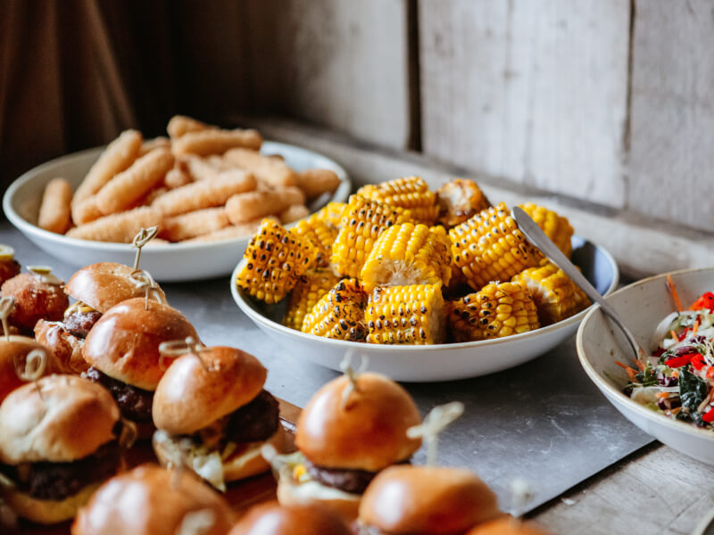 A table is full of plates of small burgers, grilled corn on the cob and bowls of salad