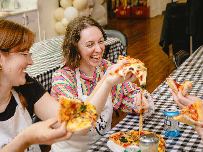 Two women smiling while eating pizza in a cooking class.