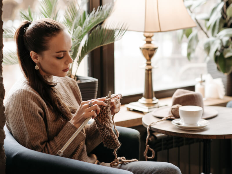 A woman is sitting in a cafe, knitting using wooden knitting needles and beige yarn