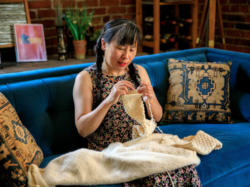 A woman is sitting on a blue sofa crocheting with cream yarn 