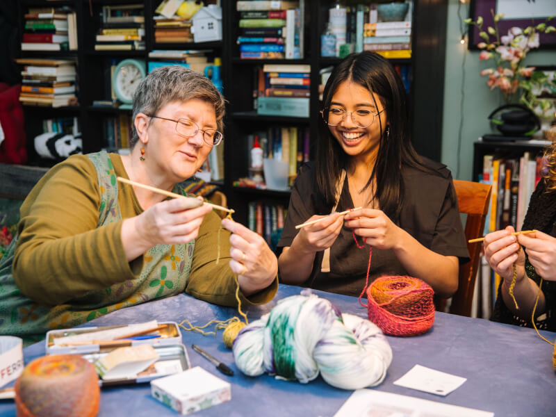 An older woman is sitting next to a younger woman at a table, teaching her how to crochet