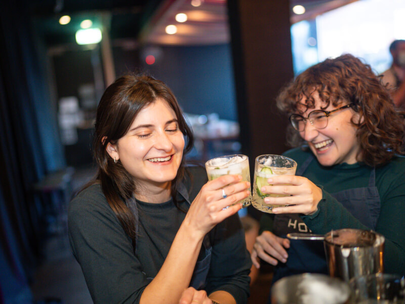 Two women are clinking glasses filled with DIY cocktails and cucumber garnishes