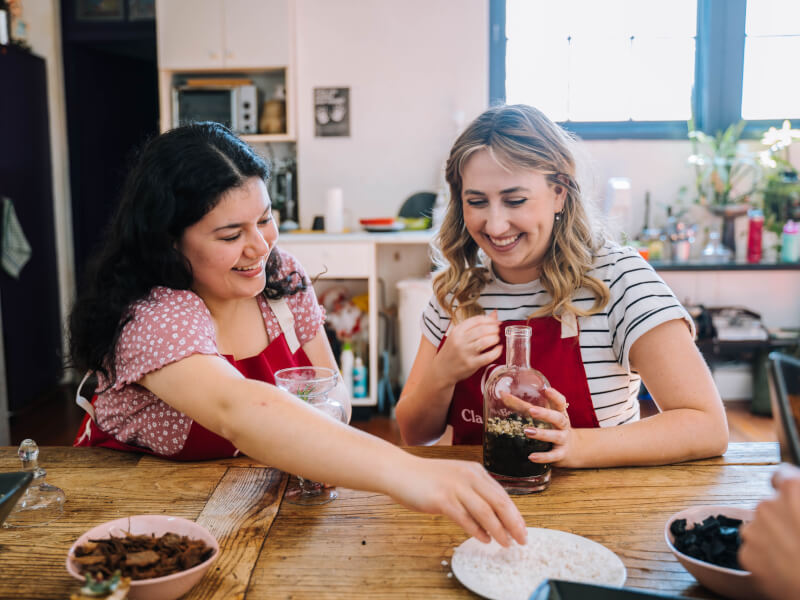 Two women wearing red aprons are sitting at a wooden table, making glass terrariums