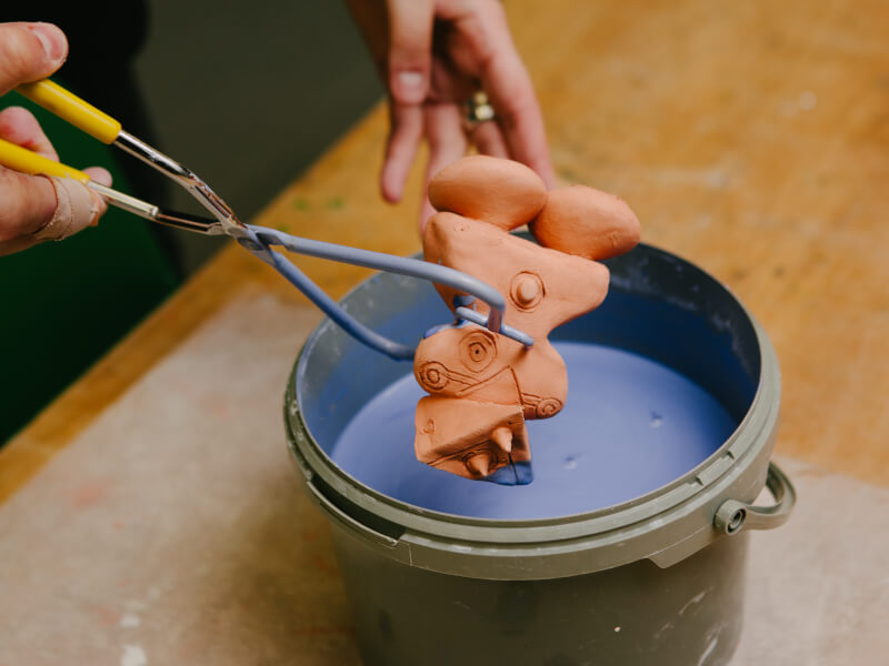 A person is holding a piece of pottery with tongs and dipping it into purple glaze