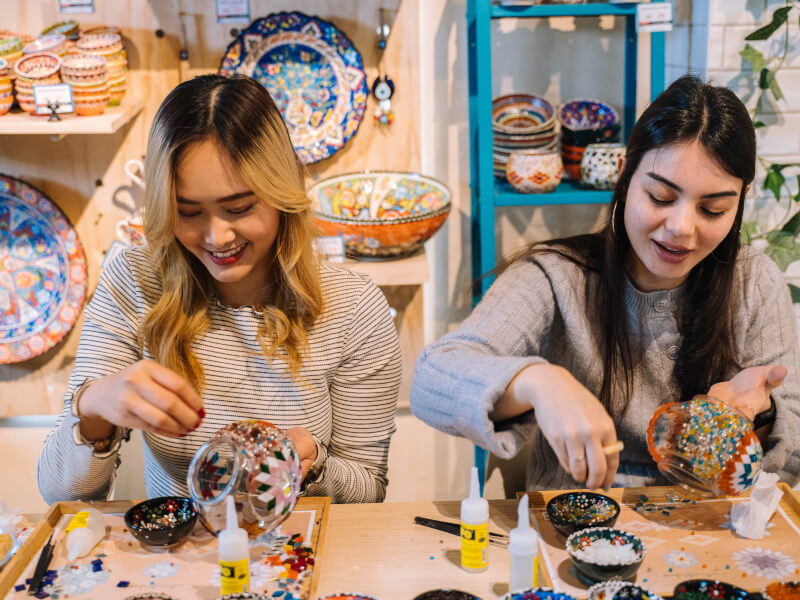 Two women sit at a table, making mosaic lampshades