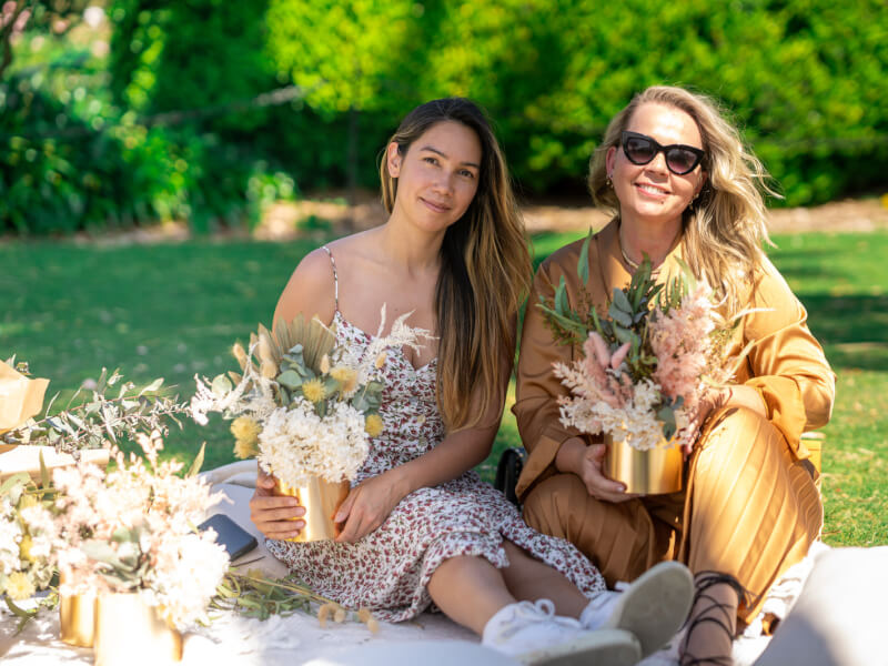 Two women are sitting on a blanket in the sunshine, holding handmade floral arrangements in gold vases