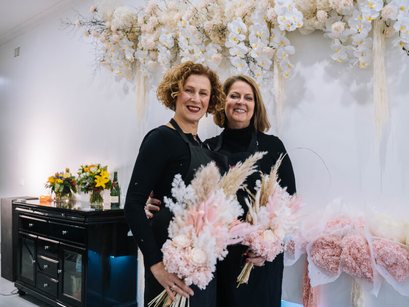 Two women are standing holding dried pink flower arrangements