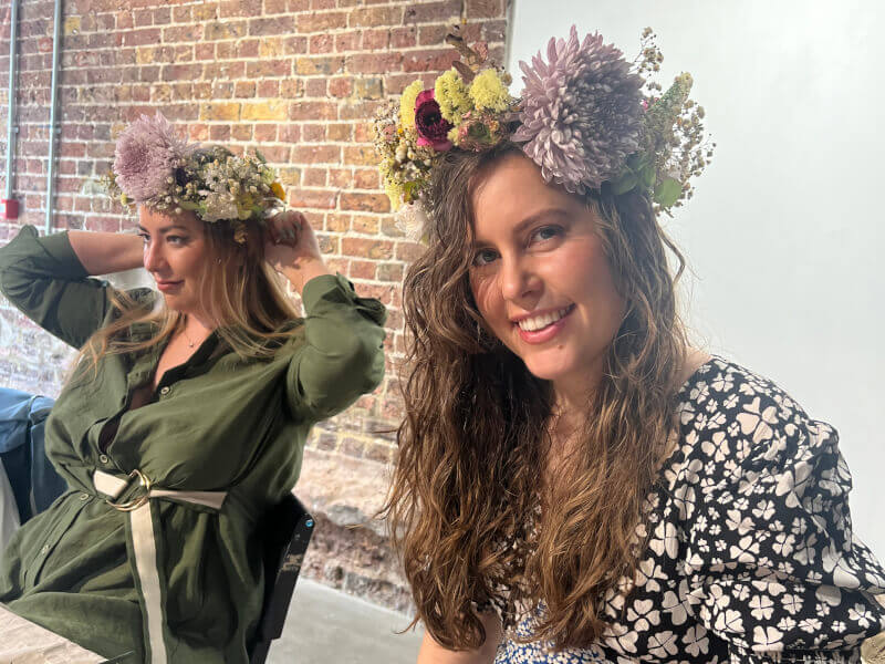 Two women sit at a table, wearing handmade flower crowns