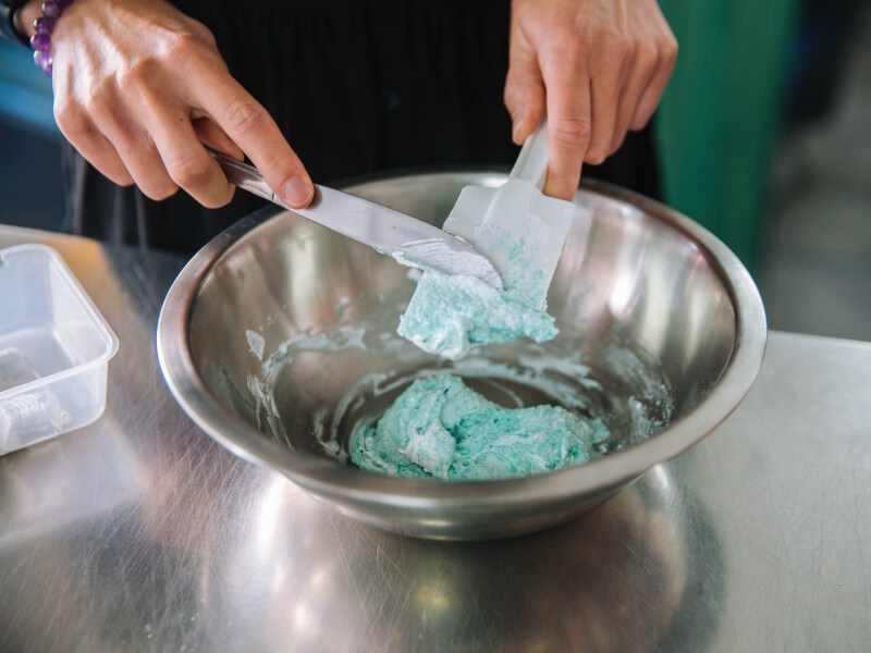 A person is scraping blue dough off of a spatula into a bowl