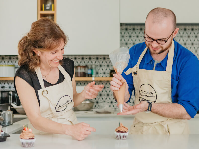 A woman is showing a man how to decorate a cupcake with chocolate frosting in a piping bag