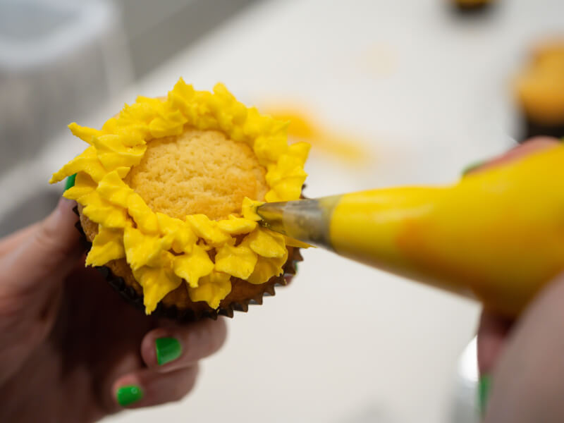 A close up of a person icing a cupcake with bright yellow frosting