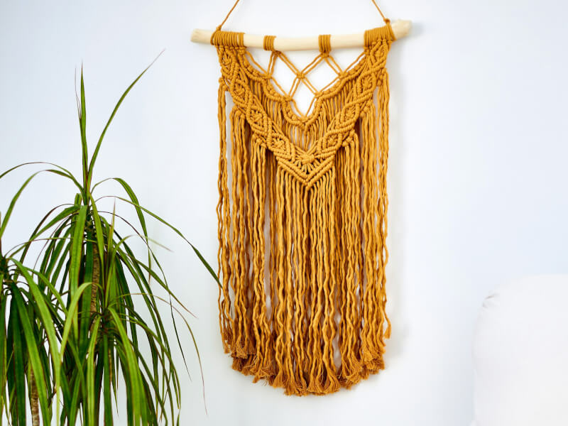 A mustard coloured macrame wall hanging hangs on a white wall with a plant to the left of it